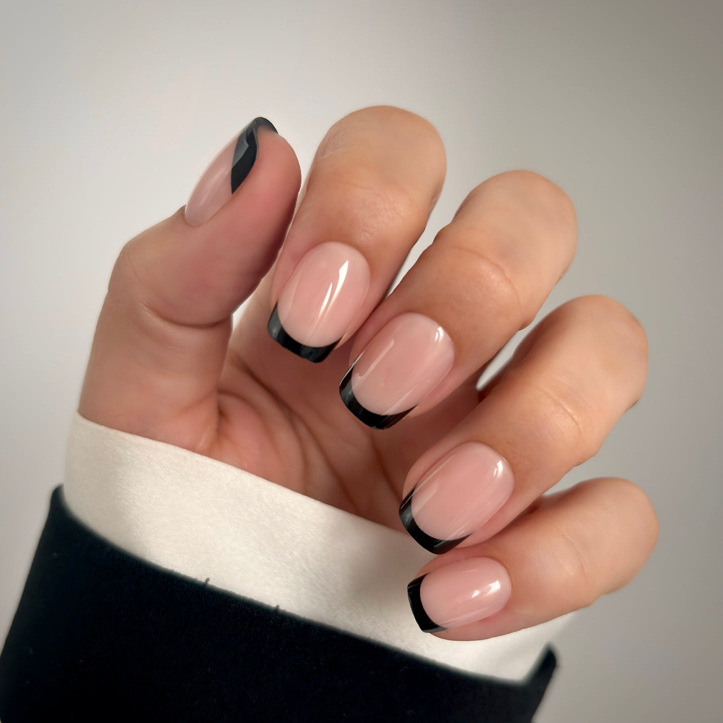A hand with medium square press-on nails featuring a bold black French tip on a soft nude base, resting against a white satin fabric in soft natural light — modern, editorial and effortlessly striking