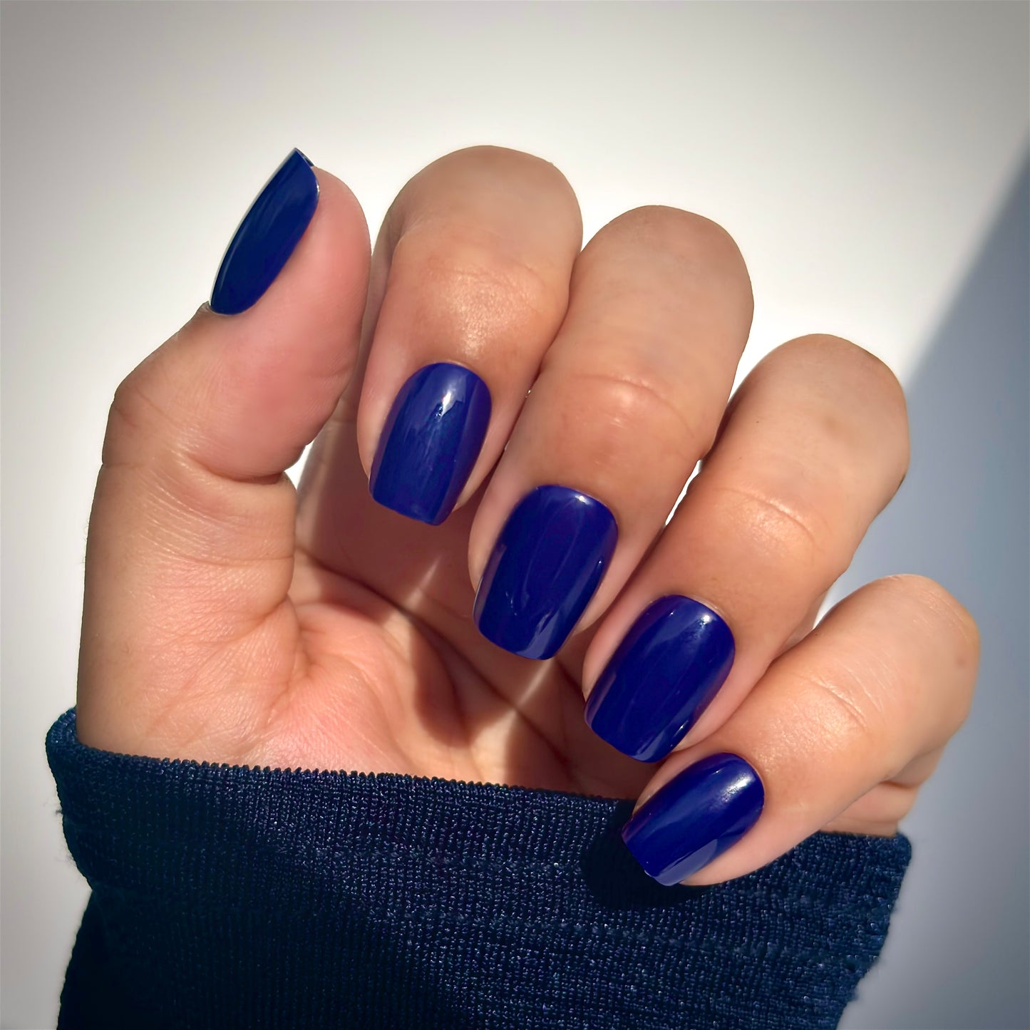A hand with medium square press-on nails in a deep, vivid navy blue, resting against a dark navy sleeve in soft natural light — bold, rich and effortlessly sophisticated with a high-gloss gel finish