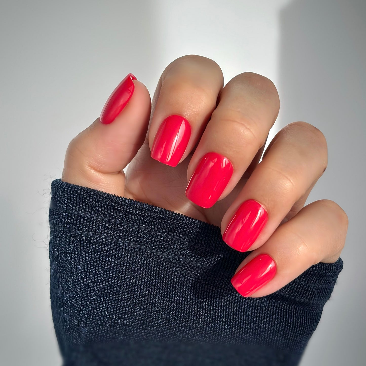 A hand with medium square press-on nails in a vivid, juicy berry red with raspberry undertones, resting against a dark knit sleeve in soft natural light — vibrant, playful and full of confidence with a high-gloss gel finish.