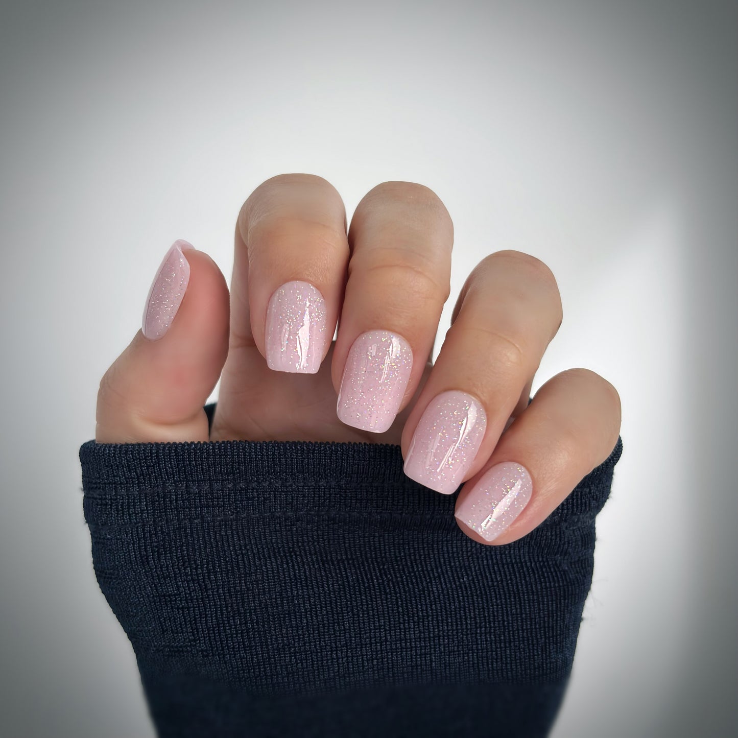 A hand with medium square press-on nails in a soft blush pink dusted with the finest shimmer — like tiny particles of diamond dust catching the light — resting against a dark navy sleeve in soft natural light, delicate, luminous and utterly feminine