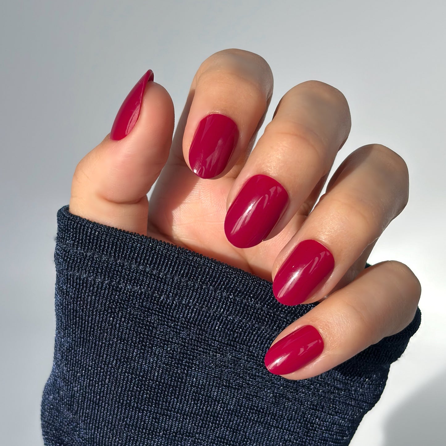 A hand with medium round press-on nails in a deep, glossy crimson red with a slight purple undertone, resting against a dark knit sleeve — rich, bold and effortlessly glamorous with a flawless gel finish