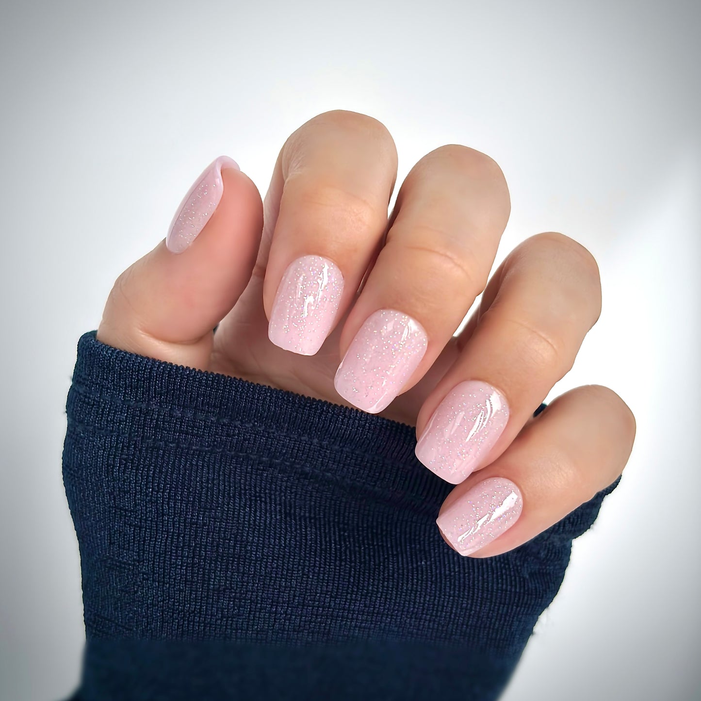 A hand with medium square press-on nails in a soft blush pink dusted with the finest shimmer — like tiny particles of diamond dust catching the light — resting against a dark navy sleeve in soft natural light, delicate, luminous and utterly feminine
