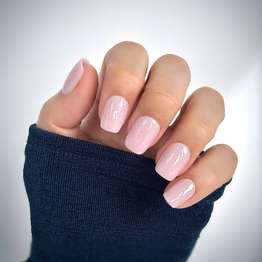 A hand with medium square press-on nails in a soft blush pink dusted with the finest shimmer — like tiny particles of diamond dust catching the light — resting against a dark navy sleeve in soft natural light, delicate, luminous and utterly feminine