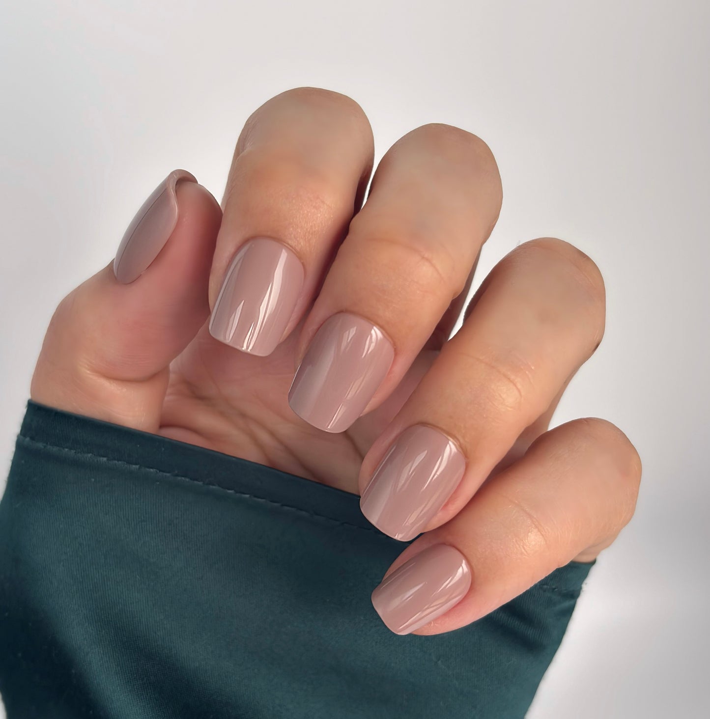 A hand with medium square press-on nails in a warm, creamy taupe nude, resting against a emerald green satin sleeve in soft natural light — understated, sophisticated and effortlessly wearable with a flawless gel finish