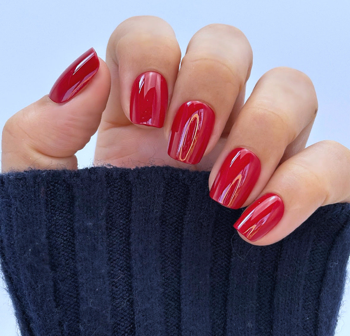 A hand with medium square press-on nails in a deep, glossy cherry red with a mirror-like glazed finish, photographed against a dark background — bold, luxurious and impossibly striking