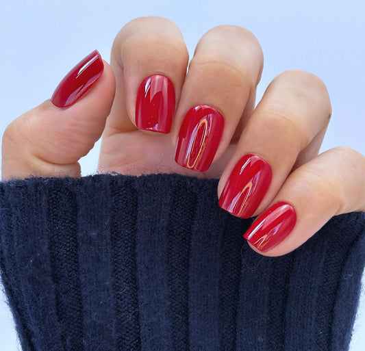 A hand with medium square press-on nails in a deep, glossy cherry red with a mirror-like glazed finish, photographed against a dark background — bold, luxurious and impossibly striking