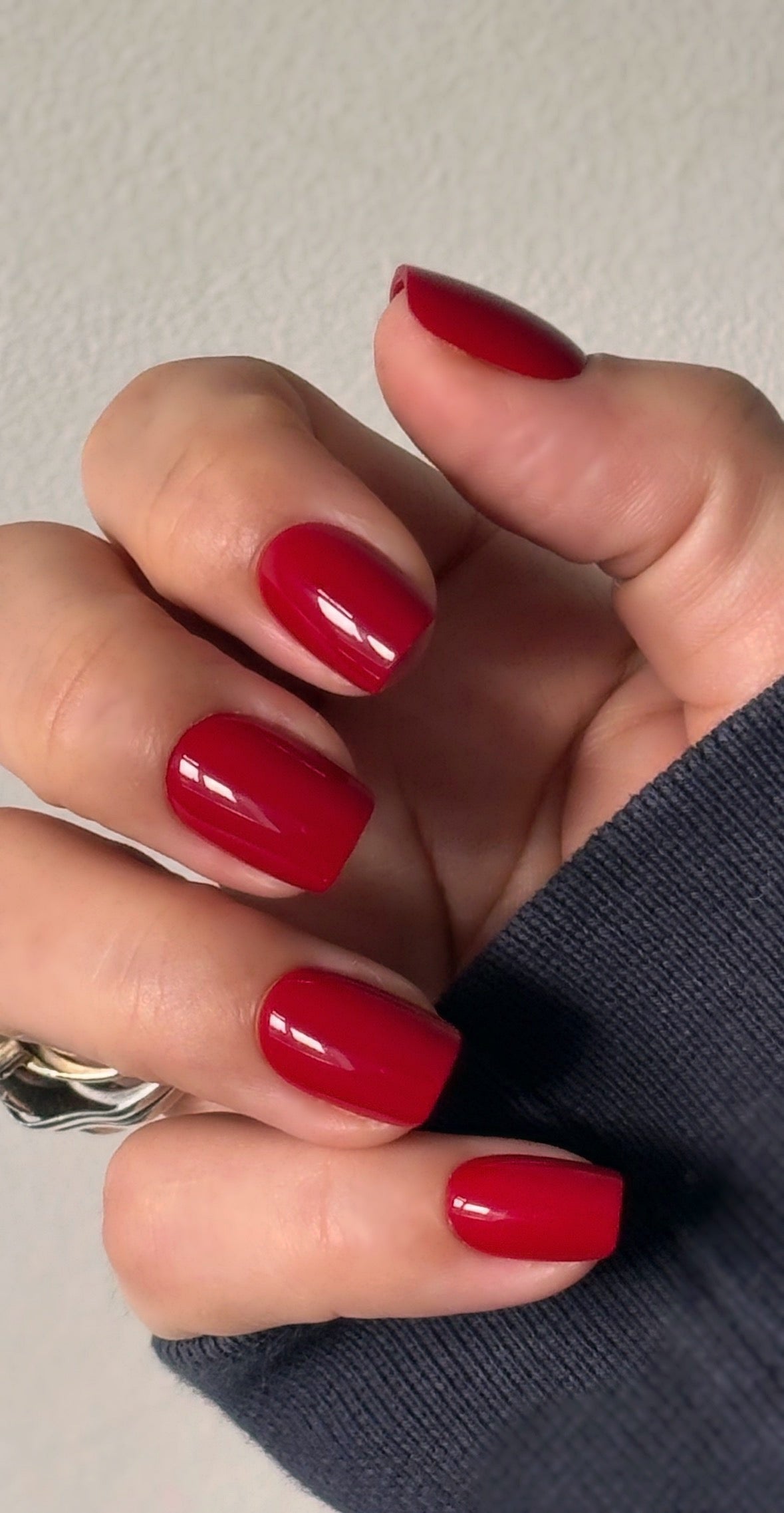 A hand with medium square press-on nails in a deep, glossy cherry red with a mirror-like glazed finish, photographed against a dark background — bold, luxurious and impossibly striking