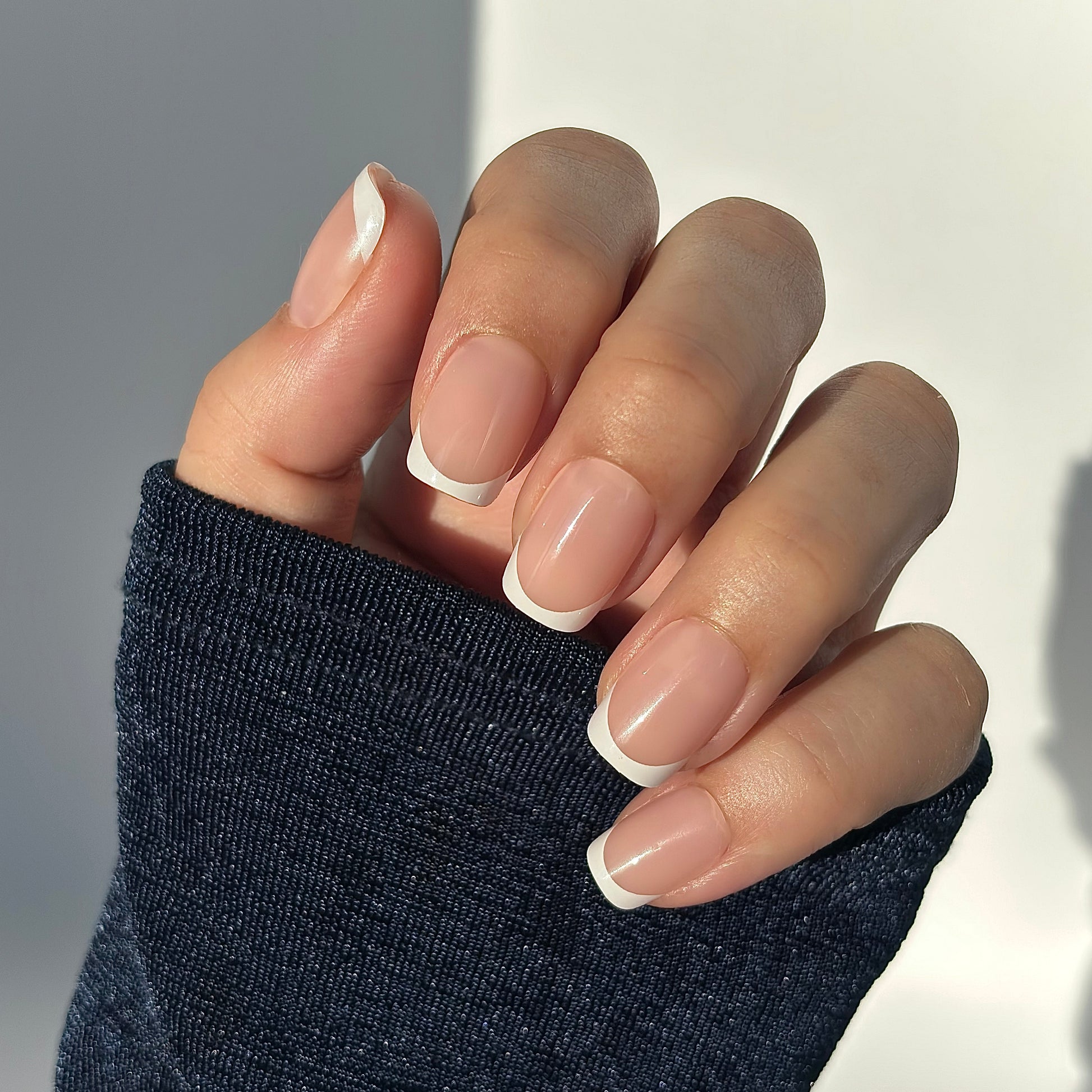 A hand with medium square press-on nails featuring a classic French tip — crisp white tips on a soft nude base — draped against a dark knit sleeve, bathed in warm golden natural light that falls softly across the hand, highlighting every curve with a gentle glow. Also available in round, extra short and almond shapes for your perfect fit.