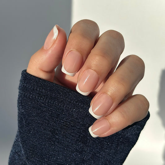 A hand with medium square press-on nails featuring a classic French tip — crisp white tips on a soft nude base — draped against a dark knit sleeve, bathed in warm golden natural light that falls softly across the hand, highlighting every curve with a gentle glow. Also available in round, extra short and almond shapes for your perfect fit.