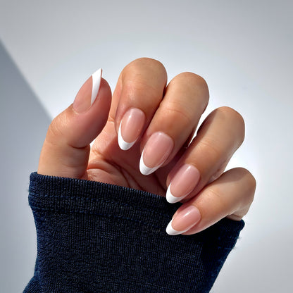 A hand with medium almond press-on nails in a classic French tip — crisp white tips on a soft nude base — elegantly posed against a dark navy sleeve, bathed in cool natural light with soft shadows that highlight every curve of the nail. Also available in extra short  round medium round and square shapes for your perfect fit.