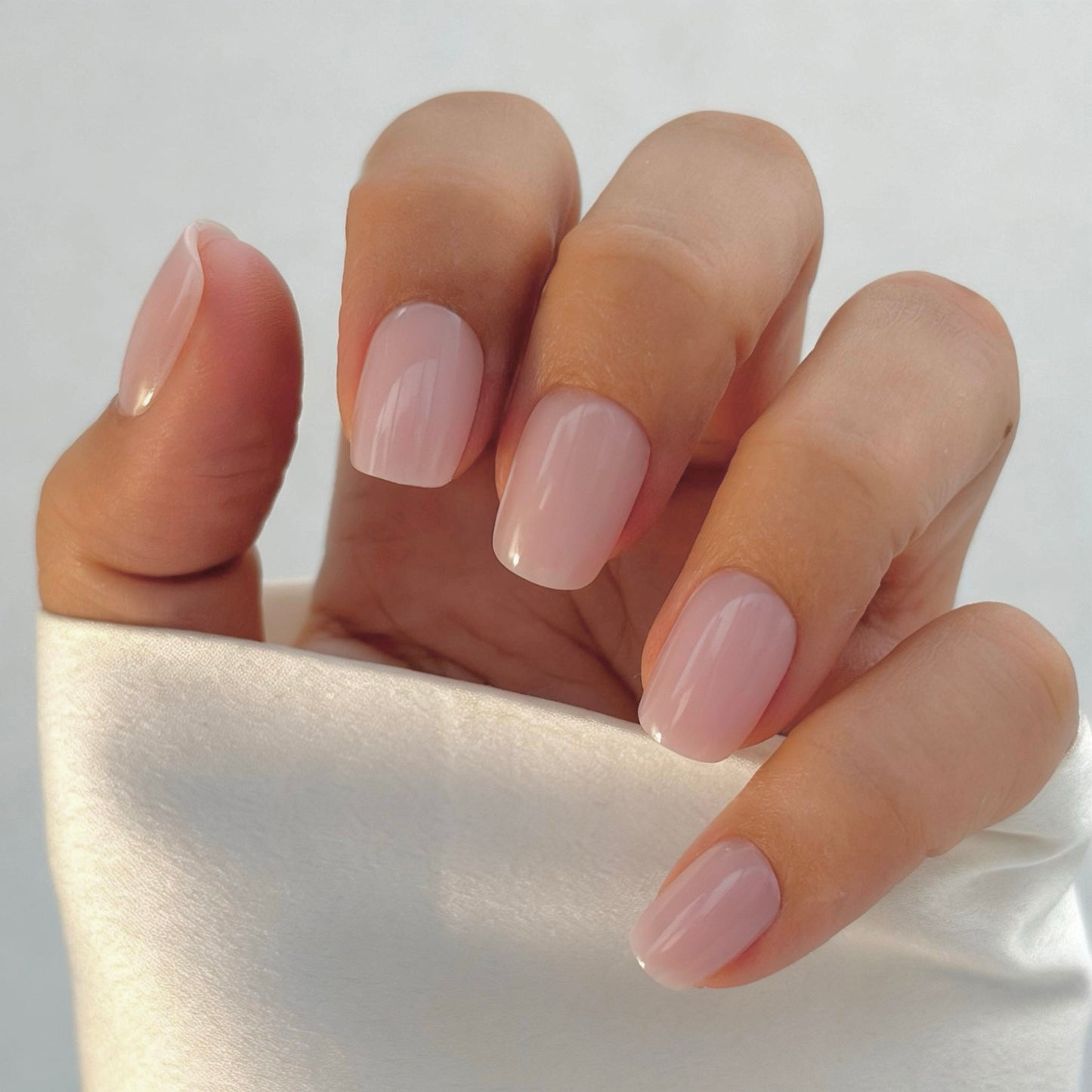 A hand with medium square press-on nails in a soft, sheer blush pink, resting against a white satin surface in natural light — delicate, feminine and effortlessly clean girl chic with a flawless gel finish