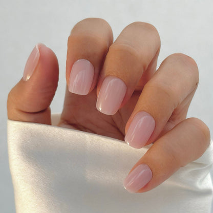 A hand with medium square press-on nails in a soft, sheer blush pink, resting against a white satin surface in natural light — delicate, feminine and effortlessly clean girl chic with a flawless gel finish