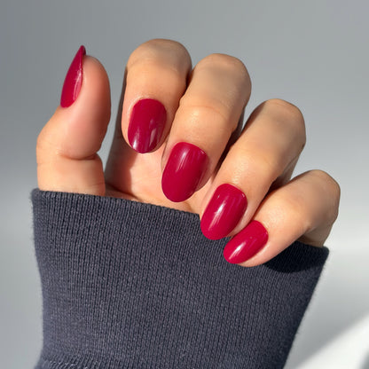 A hand with medium round press-on nails in a deep, glossy crimson red with a slight purple undertone, resting against a dark knit sleeve — rich, bold and effortlessly glamorous with a flawless gel finish
