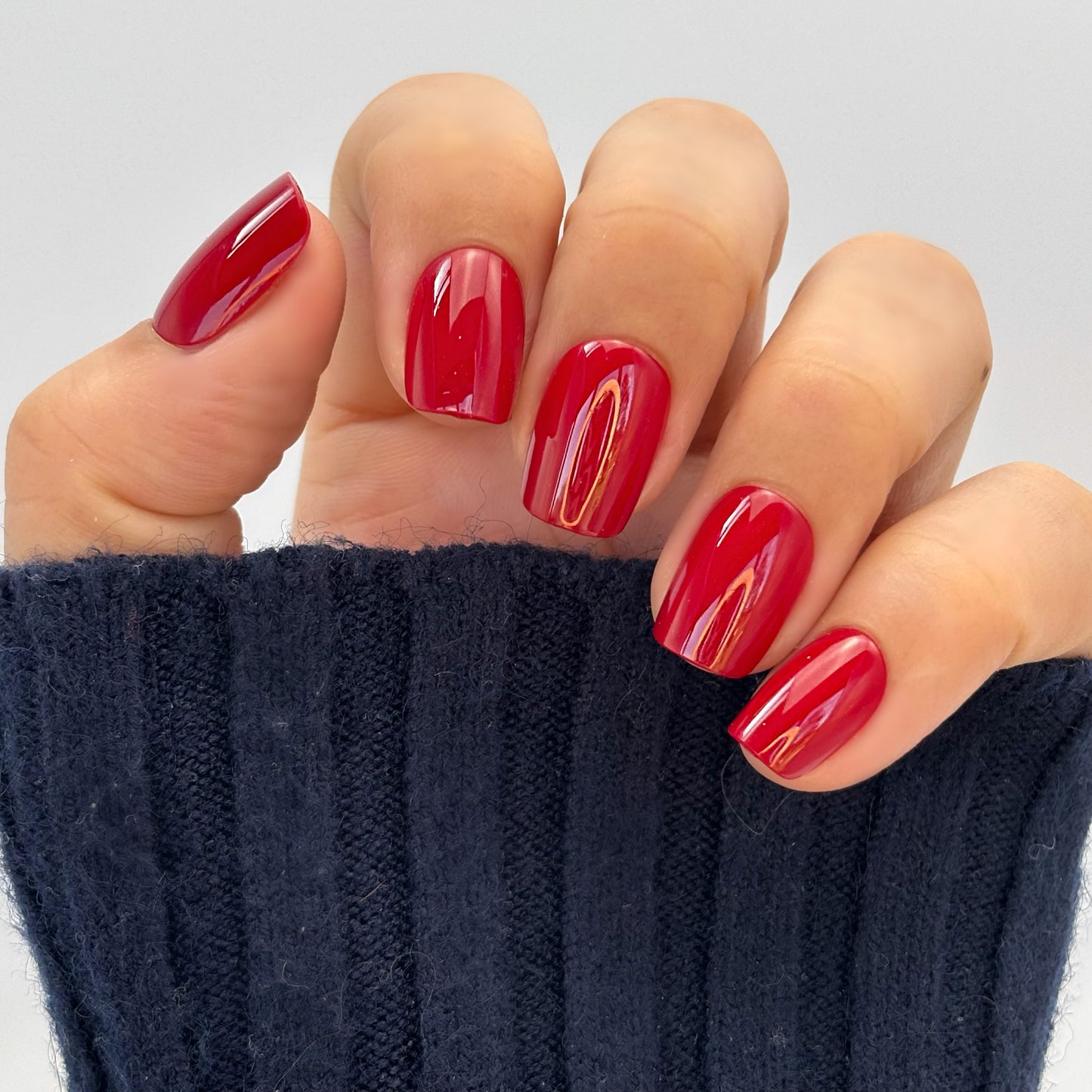 A hand with medium square press-on nails in a deep, glossy cherry red with a mirror-like glazed finish, photographed against a dark background — bold, luxurious and impossibly striking