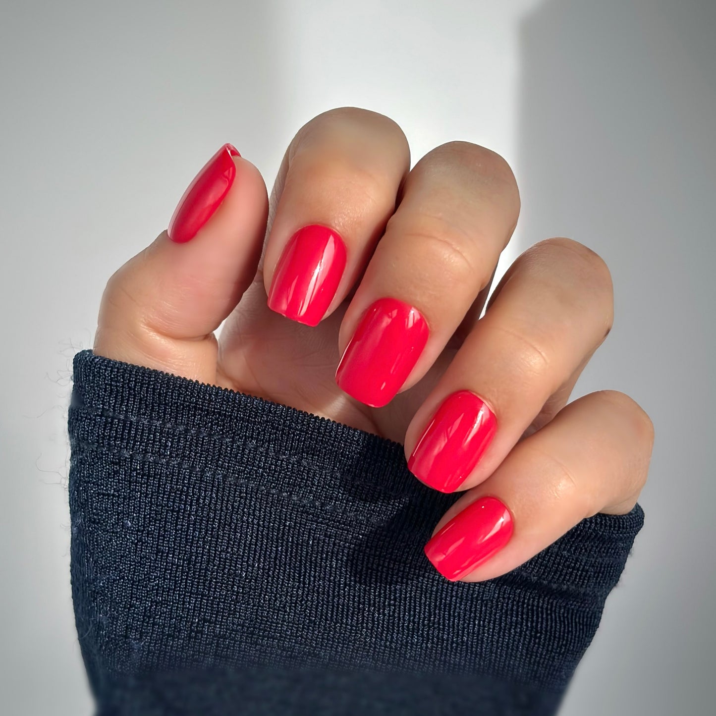 A hand with medium square press-on nails in a vivid, juicy berry red with raspberry undertones, resting against a dark knit sleeve in soft natural light — vibrant, playful and full of confidence with a high-gloss gel finish.