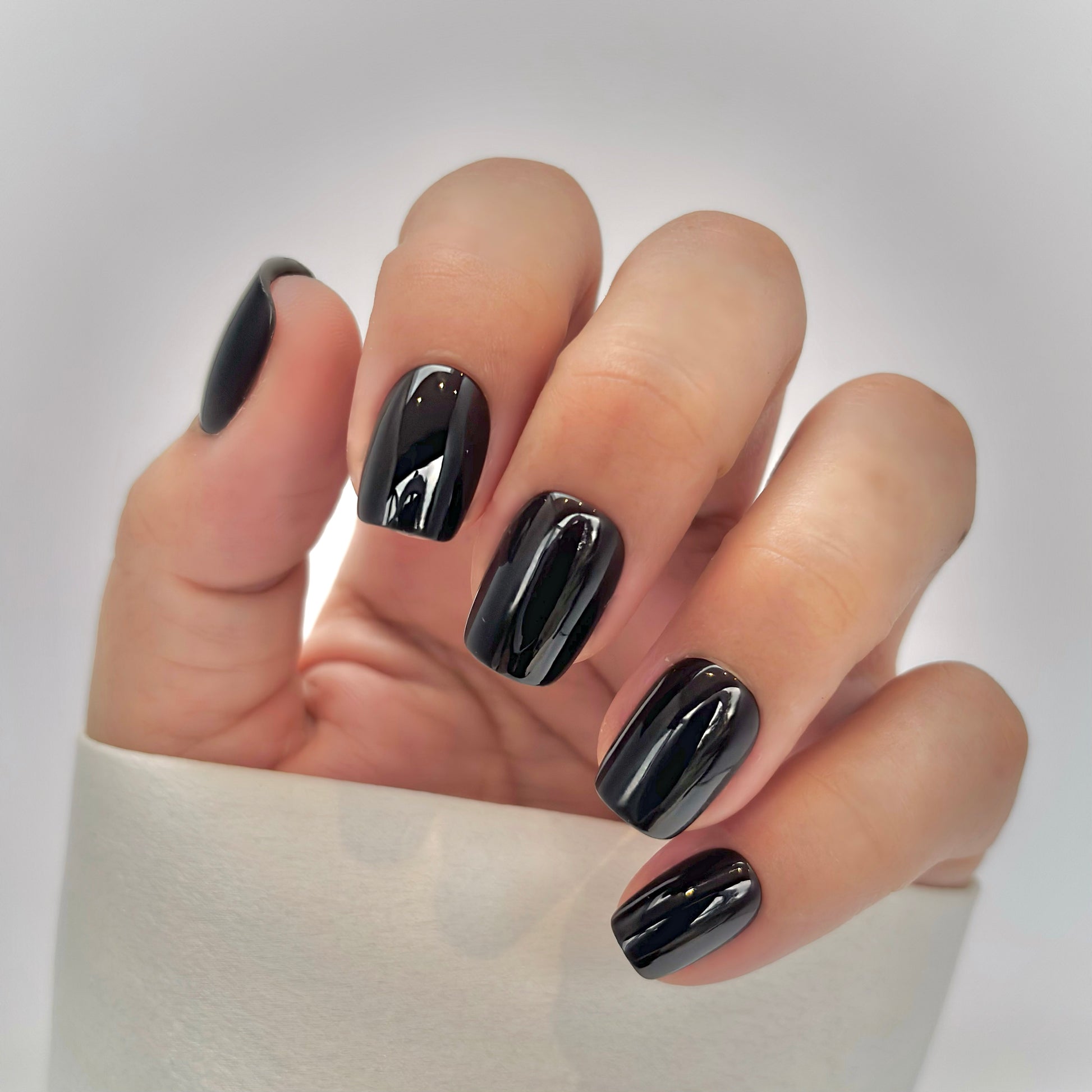 A hand with medium square press-on nails in a deep, opulent dark brown with a mirror-like high-gloss finish, resting against a cream satin sleeve, bathed in gentle natural light with soft shadows that fall across the background — rich, luxurious and effortlessly sophisticated