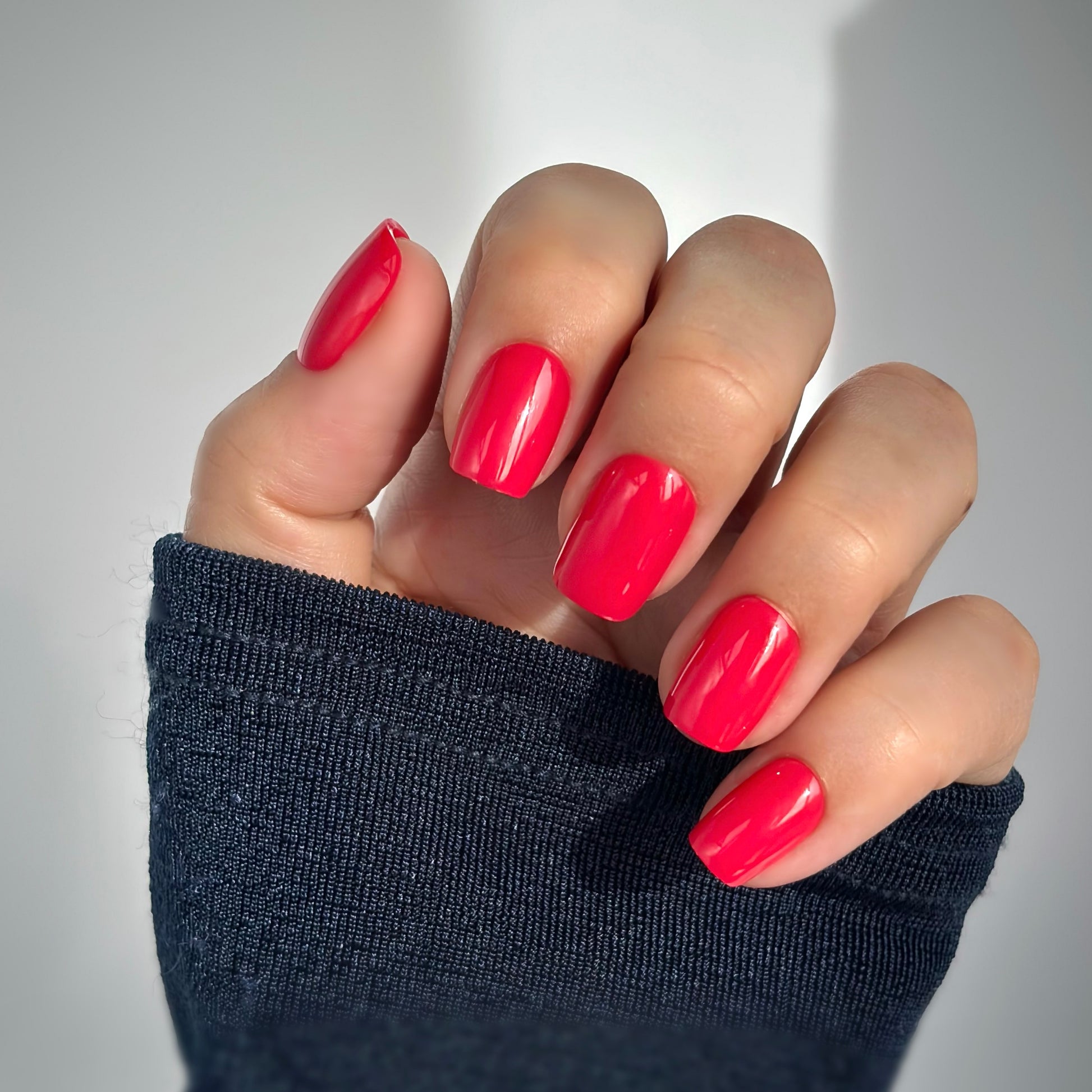 A hand with medium square press-on nails in a vivid, juicy berry red with raspberry undertones, resting against a dark knit sleeve in soft natural light — vibrant, playful and full of confidence with a high-gloss gel finish.