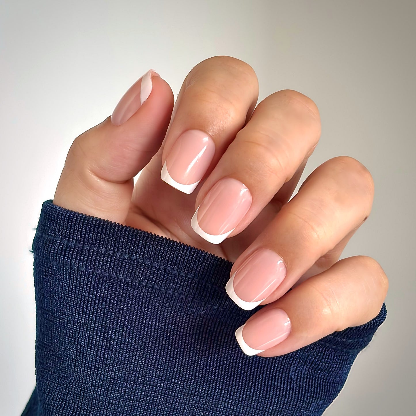 A hand with medium square press-on nails featuring a classic French tip — crisp white tips on a soft nude base — draped against a dark knit sleeve. Also available in round, extra short and almond shapes for your perfect fit.