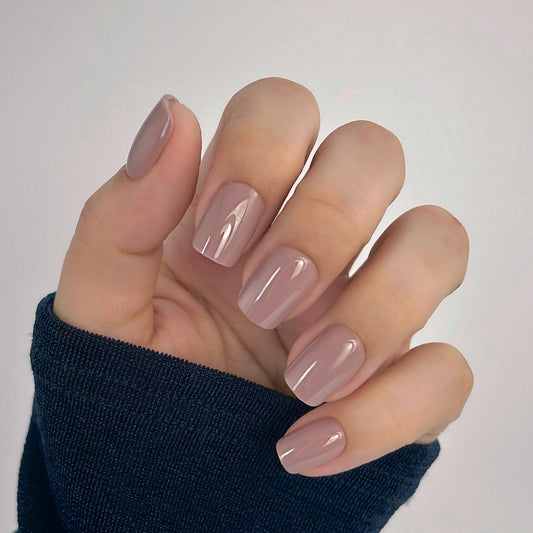 A hand with medium square press-on nails in a warm, creamy taupe nude, resting against a dark navy sleeve in soft natural light — understated, sophisticated and effortlessly wearable with a flawless gel finish