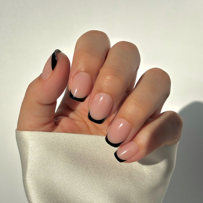 A hand with medium square press-on nails featuring a bold black French tip on a soft nude base, resting against a white satin fabric in soft natural light — modern, editorial and effortlessly striking