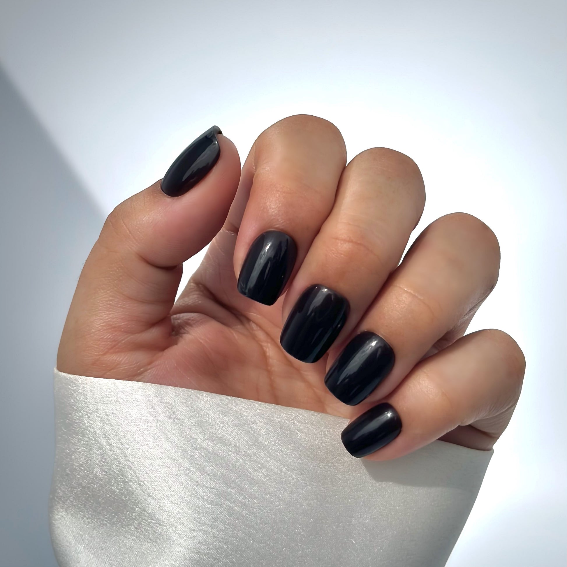 A hand with medium square press-on nails in a deep, opulent dark brown with a mirror-like high-gloss finish, resting against a cream satin sleeve, bathed in gentle natural light with soft shadows that fall across the background — rich, luxurious and effortlessly sophisticated