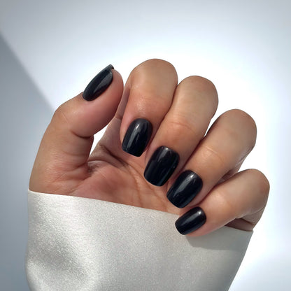 A hand with medium square press-on nails in a deep, opulent dark brown with a mirror-like high-gloss finish, resting against a cream satin sleeve, bathed in gentle natural light with soft shadows that fall across the background — rich, luxurious and effortlessly sophisticated
