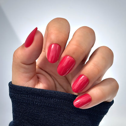 A hand with medium round press-on nails in a rich, glossy cherry red, photographed in natural light with soft shadows — bold, timeless and effortlessly glamorous with a mirror-like gel finish. Also available in an extra short style as Cherry Rouge Petite