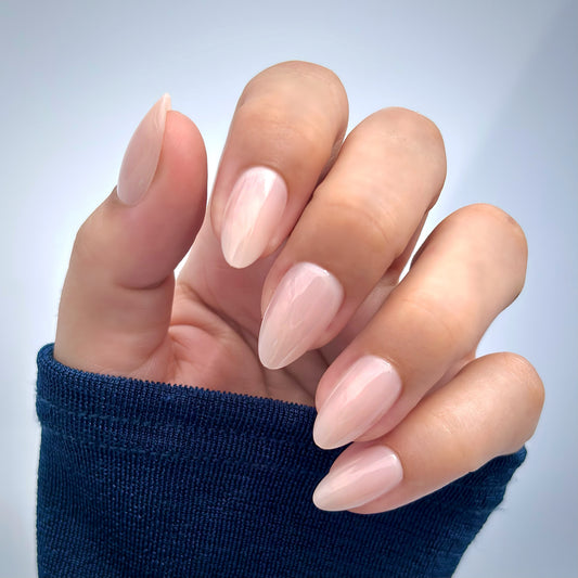 A hand with medium almond press-on nails in a soft, creamy ivory pink, elegantly posed against a navy sleeve — understated, refined and effortlessly sophisticated with a flawless gel finish