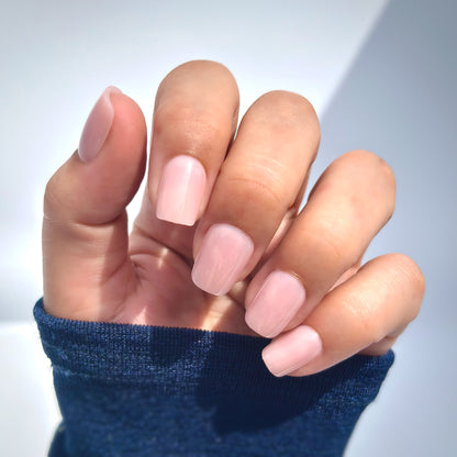 A hand with medium square press-on nails in a soft, muted blush pink, resting gently against a striking blue fabric in a natural glistening  light with a white background showing gentle shadows— delicate, romantic and bridal-ready with a flawless gel finish