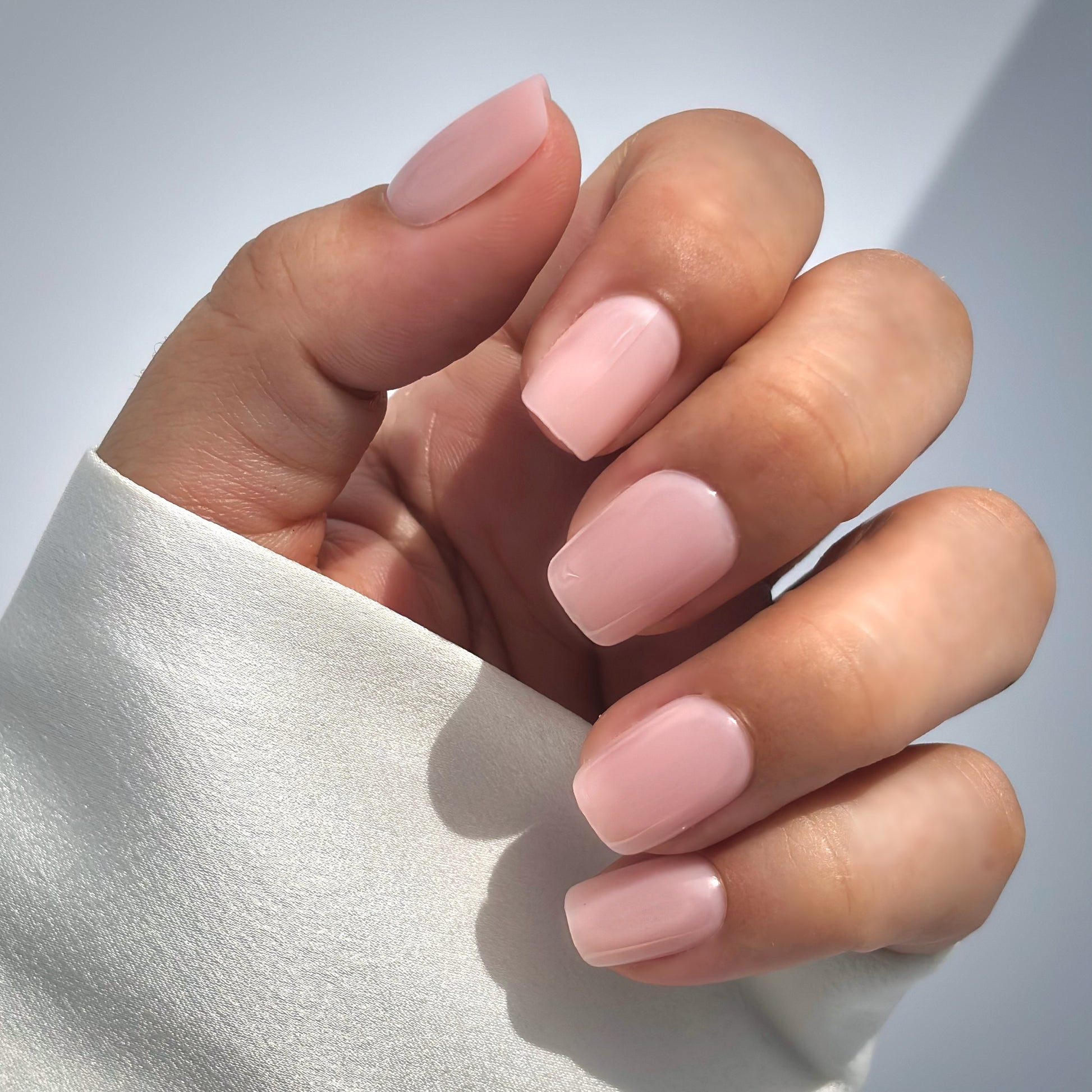 A hand with medium square press-on nails in a soft, muted blush pink, resting gently against a white satin fabric in a natural glistening  light with a white background — delicate, romantic and bridal-ready with a flawless gel finish
