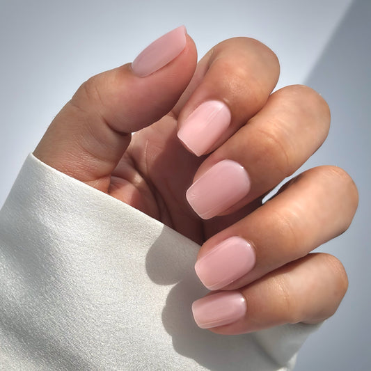 A hand with medium square press-on nails in a soft, muted blush pink, resting gently against a white satin fabric in a natural glistening  light with a white background — delicate, romantic and bridal-ready with a flawless gel finish