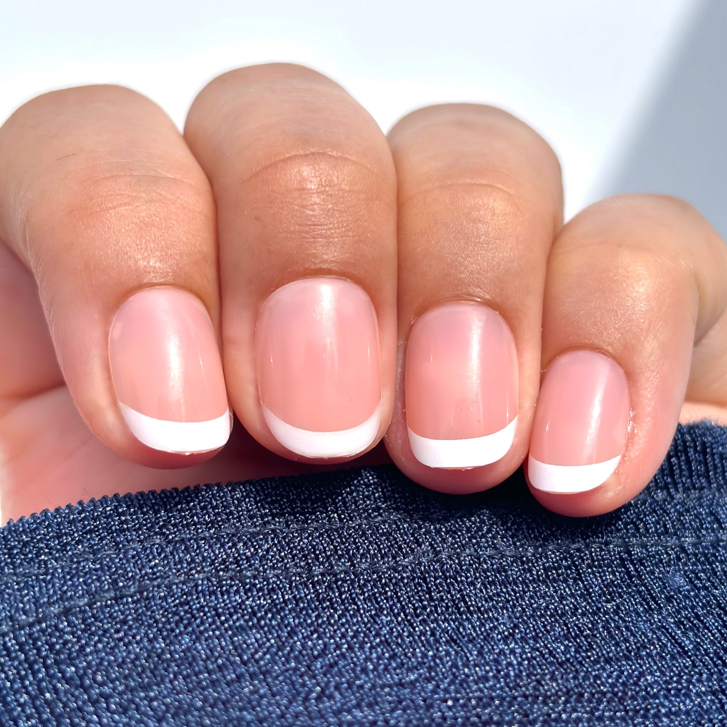 A close up of a hand with extra short round French tip press-on nails — a classic white tip on a soft nude base — draped against a navy sleeve in natural light, clean, understated and effortlessly polished