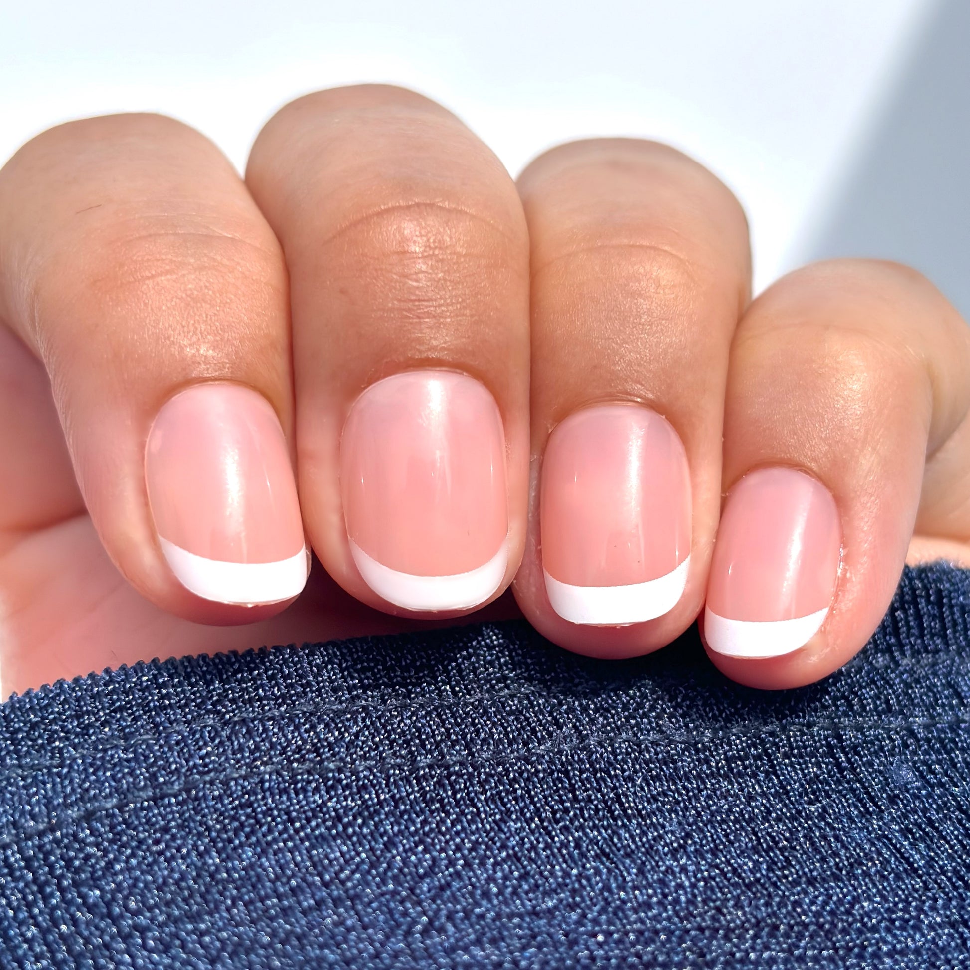 A close up of a hand with extra short round French tip press-on nails — a classic white tip on a soft nude base — draped against a navy sleeve in natural light, clean, understated and effortlessly polished