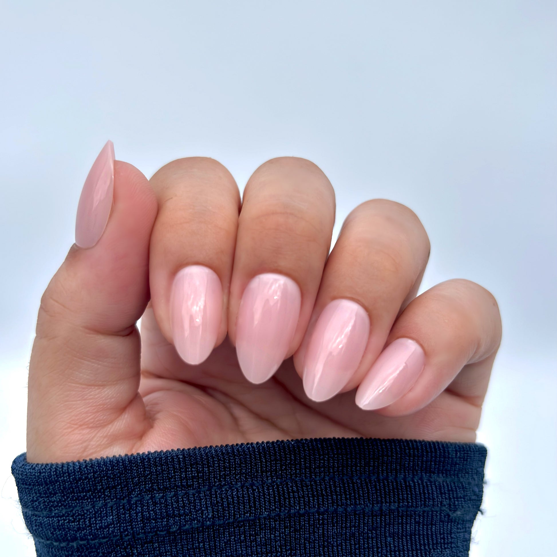 A hand adorned with soft blush pink press-on nails in a medium almond shape, elegantly posed against a blue sleeve — high-gloss gel finish, feminine and salon-perfect