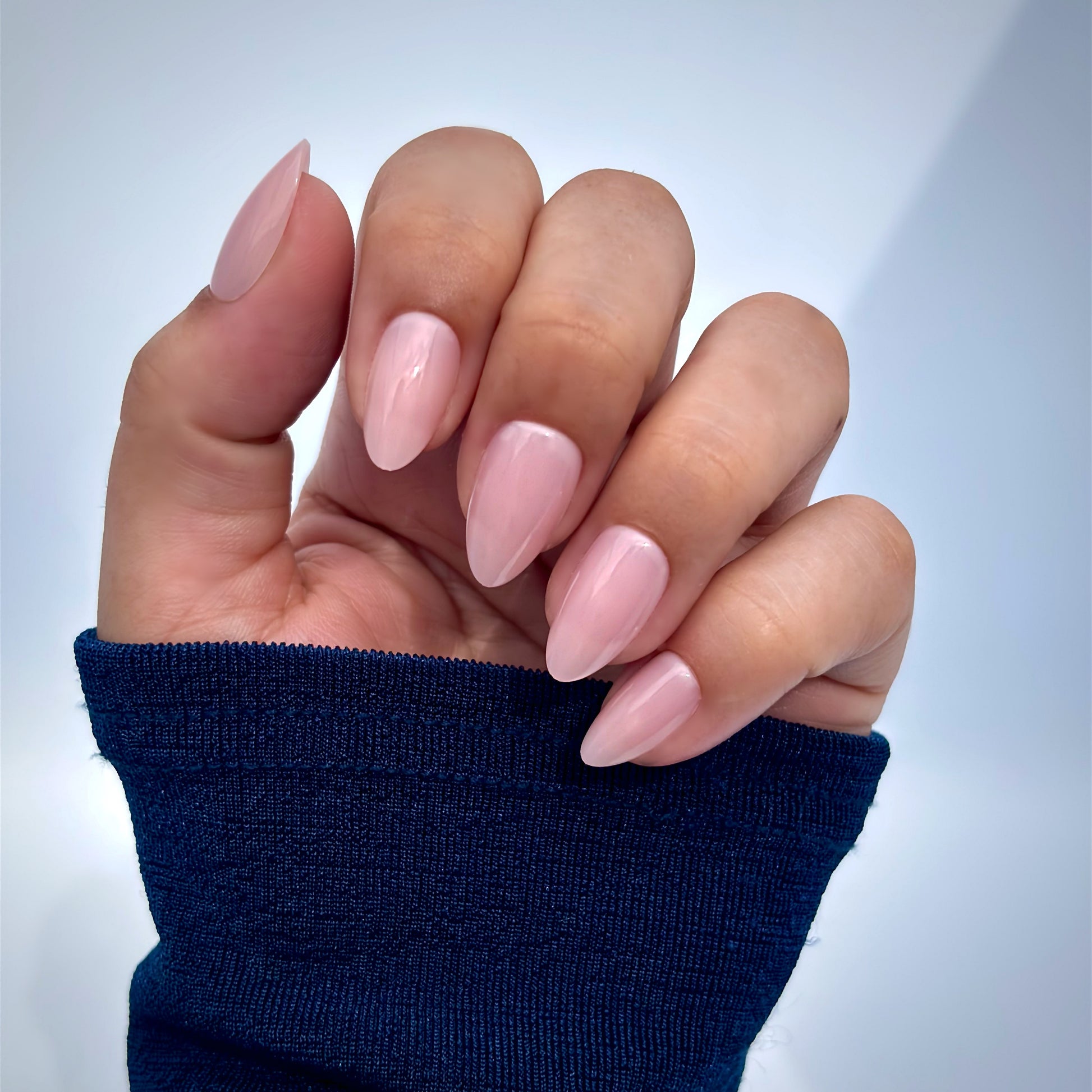 A hand adorned with soft blush pink press-on nails in a medium almond shape, elegantly posed against a blue sleeve — high-gloss gel finish, feminine and salon-perfect