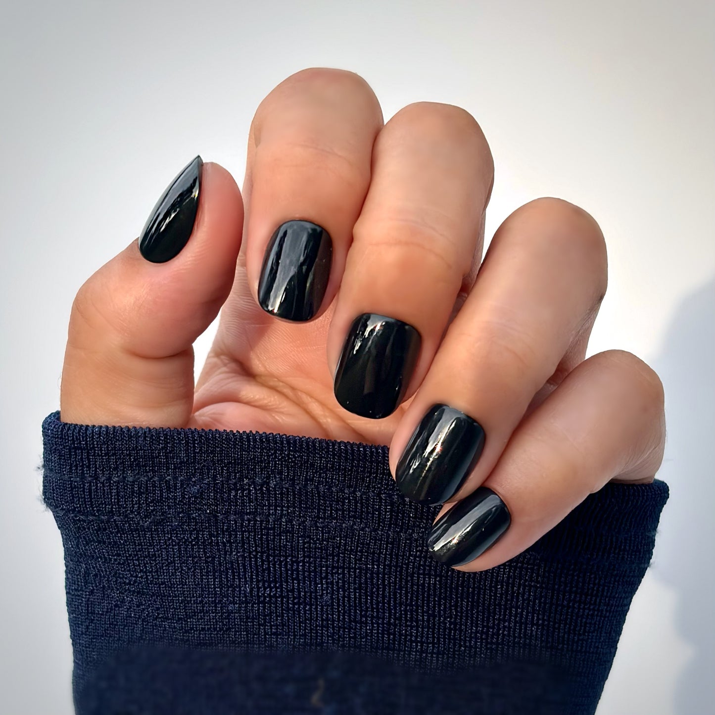 A hand with extra short round press-on nails in a deep, glossy black, resting against a navy blue sleeve — bold, sleek and impossibly chic