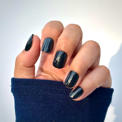 A hand with extra short round press-on nails in a deep, glossy black, resting against a navy blue sleeve — bold, sleek and impossibly chic