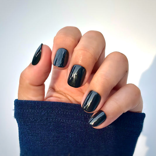 A hand with extra short round press-on nails in a deep, glossy black, resting against a navy blue sleeve — bold, sleek and impossibly chic