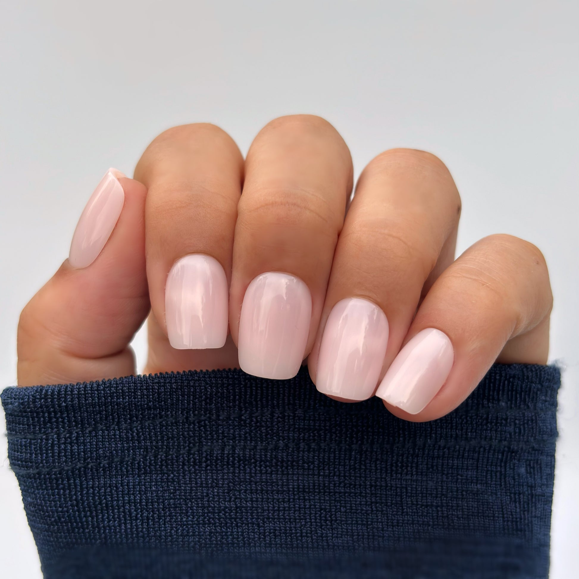A hand with medium square press-on nails in a soft milky pink, resting gently against a navy blue sleeve — clean, minimal and effortlessly polished