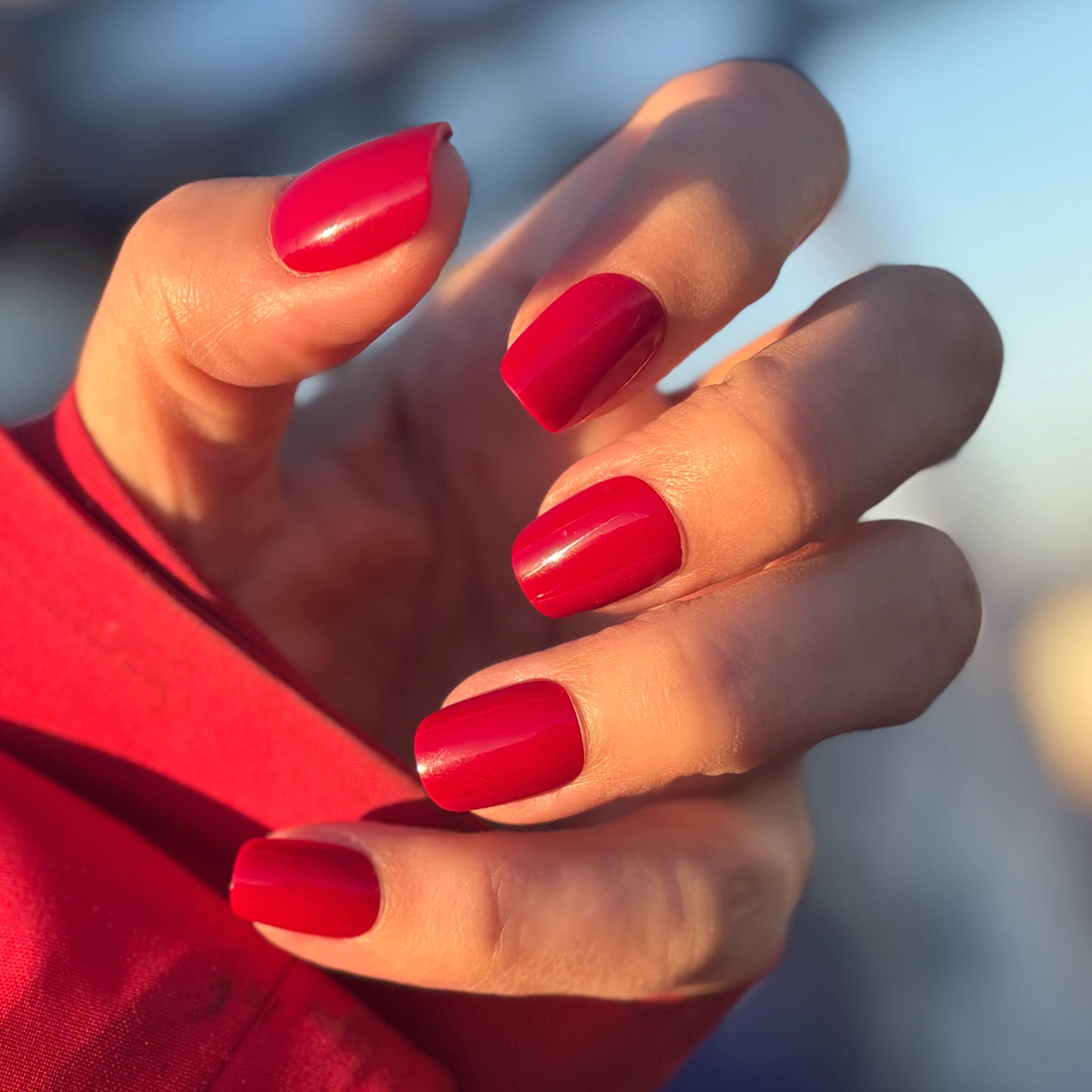 A hand with medium square press-on nails in a deep, glossy cherry red with a mirror-like glazed finish, photographed against a dark background — bold, luxurious and impossibly striking